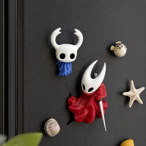 May include: A collection of decorative magnets featuring a white horned character with a blue cape, a white horned character with a red cape and a needle, a small beige pufferfish, a white starfish, and a small tan seashell on a dark surface.