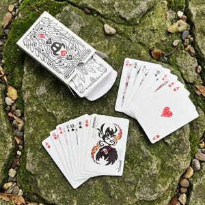 May include: A deck of playing cards with a white box featuring a black and white design. Several cards are fanned out, displaying suits and numbers. The Joker card has a unique illustration with the word "Joker" on it.