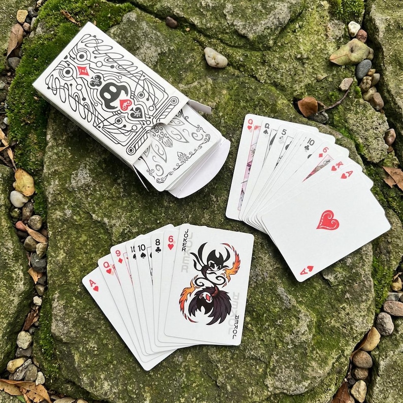 May include: A deck of playing cards with a white box featuring a black and white design. Several cards are fanned out, displaying suits and numbers. The Joker card has a unique illustration with the word "Joker" on it.