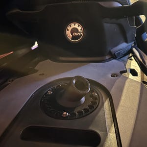 May include: Close-up of a black snowmobile component featuring a circular fuel filler neck and a black seat with the BRP logo. The image highlights the machine's design and mechanical details.