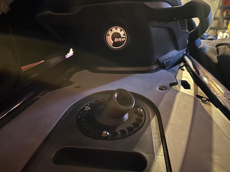 K&ouml;nnte beinhalten: Nahaufnahme einer schwarzen Schneemobilkomponente mit einem kreisf&ouml;rmigen Kraftstoffeinf&uuml;llstutzen und einem schwarzen Sitz mit dem BRP-Logo. Das Bild zeigt das Design und die mechanischen Details der Maschine.