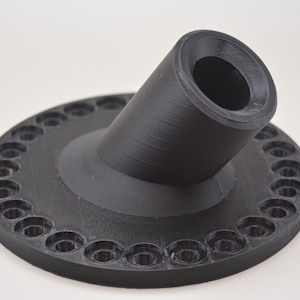 K&ouml;nnte beinhalten: Ein schwarzes, 3D-gedrucktes Objekt mit kreisf&ouml;rmiger Basis und abgewinkeltem, zylindrischem Bestandteil. Der Sockel weist gleichm&auml;&szlig;ig verteilte L&ouml;cher am Umfang auf. Das Objekt besteht aus mattem, schwarzem Material und liegt auf wei&szlig;em Untergrund.