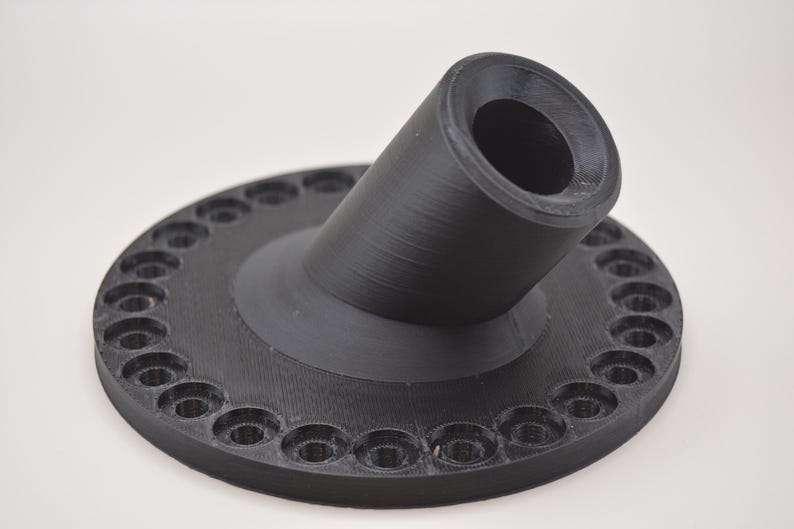 K&ouml;nnte beinhalten: Ein schwarzes, 3D-gedrucktes Objekt mit kreisf&ouml;rmiger Basis und abgewinkeltem, zylindrischem Bestandteil. Der Sockel weist gleichm&auml;&szlig;ig verteilte L&ouml;cher am Umfang auf. Das Objekt besteht aus mattem, schwarzem Material und liegt auf wei&szlig;em Untergrund.