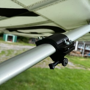 May include: A black clamp securely fastens to a silver metal pole, with a small black knob and screw visible. The clamp is attached to a white, textured surface, likely part of a larger structure. The background shows green grass and a glimpse of a building.