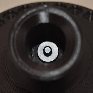 K&ouml;nnte beinhalten: Nahaufnahme eines schwarzen, kreisf&ouml;rmigen Objekts mit einer zentralen, zylindrischen &Ouml;ffnung. Eine kleine, silberne Scheibe ist in der &Ouml;ffnung zentriert. Der &auml;u&szlig;ere Rand hat eine Reihe kleiner, gleichm&auml;&szlig;ig verteilter L&ouml;cher.