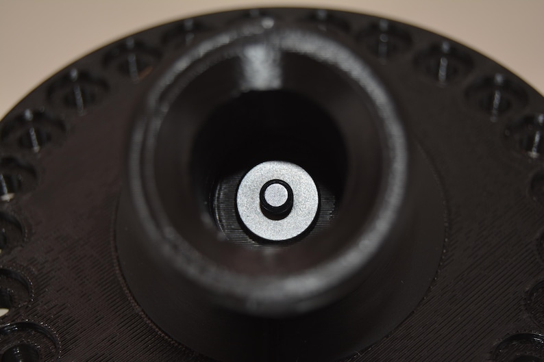 K&ouml;nnte beinhalten: Nahaufnahme eines schwarzen, kreisf&ouml;rmigen Objekts mit einer zentralen, zylindrischen &Ouml;ffnung. Eine kleine, silberne Scheibe ist in der &Ouml;ffnung zentriert. Der &auml;u&szlig;ere Rand hat eine Reihe kleiner, gleichm&auml;&szlig;ig verteilter L&ouml;cher.