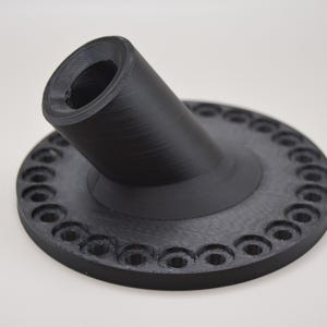 K&ouml;nnte beinhalten: Ein schwarzes, 3D-gedrucktes Objekt mit kreisf&ouml;rmiger Basis und abgewinkeltem, zylindrischem Bestandteil. Die Basis weist gleichm&auml;&szlig;ig verteilte, kreisf&ouml;rmige Vertiefungen am Umfang auf. Das Objekt besteht aus mattem, schwarzem Material.