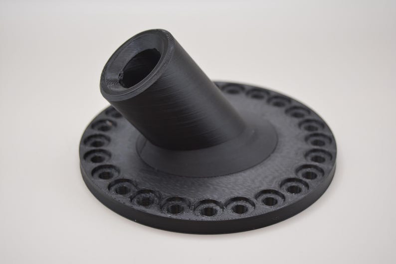 K&ouml;nnte beinhalten: Ein schwarzes, 3D-gedrucktes Objekt mit kreisf&ouml;rmiger Basis und abgewinkeltem, zylindrischem Bestandteil. Die Basis weist gleichm&auml;&szlig;ig verteilte, kreisf&ouml;rmige Vertiefungen am Umfang auf. Das Objekt besteht aus mattem, schwarzem Material.