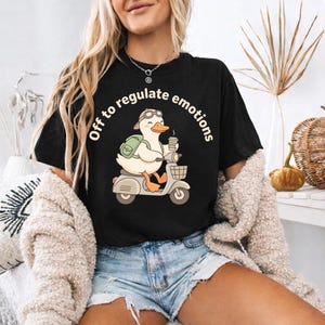 May include: Black t-shirt featuring a cartoon duck on a scooter, wearing goggles and a backpack, holding a coffee cup. The text "Off to regulate emotions" is in a circular arrangement.