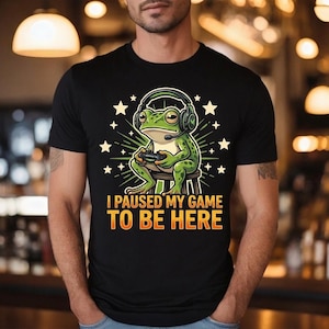 Peut inclure: T-shirt noir avec une grenouille de dessin animé portant un casque et tenant une manette de jeu. La grenouille est verte avec des détails jaunes, assise sur un tabouret, entourée d'étoiles. Le texte sur le t-shirt dit "I PAUSED MY GAME TO BE HERE."