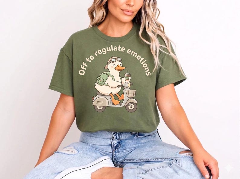 May include: Olive green t-shirt with a cartoon duck on a scooter, wearing goggles and a backpack, and holding a coffee cup. The text "Off to regulate emotions" is printed above the duck. The shirt is worn with blue jeans.
