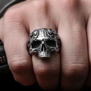 May include: A silver skull ring with intricate detailing, featuring a detailed skull design. The ring is worn on a hand, showcasing the gothic and edgy style of the jewelry. The ring is a statement piece.