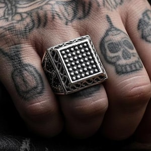 Puede incluir: Un anillo cuadrado plateado con un centro punteado y detalles laterales ornamentados. El anillo se lleva en un dedo. El fondo está borroso, con una motocicleta visible.