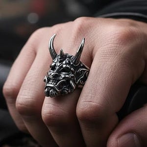 May include: A silver-colored ring featuring a detailed demon face with horns. The ring is worn on a hand, showcasing the intricate design and metallic sheen. The ring's design includes sharp features and a textured surface.