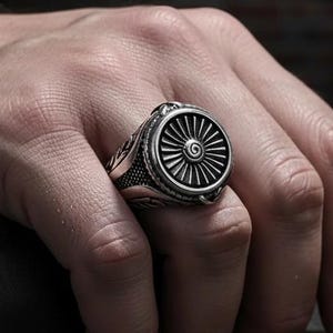 Peut inclure: Bague argentée ornée d'un motif détaillé de moteur à réaction. La bague présente une face circulaire avec des lignes radiales et une spirale centrale. La bande présente un motif gravé. La bague est portée au doigt.