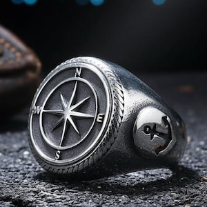 May include: A silver ring featuring a compass rose design on the top and an anchor on the side. The compass rose has the cardinal points. The ring is set on a dark, textured surface.