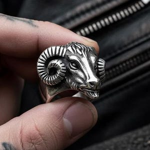 May include: A silver-colored ring featuring a detailed ram's head design. The ram's head has curved horns, a textured face, and is set on a thick band. The ring is held against a dark background, showcasing the intricate details of the jewelry.