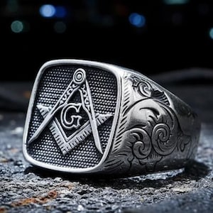 May include: A silver ring with a square face displaying the Masonic Square and Compasses emblem. The ring has ornate scrollwork on the sides. The background is dark and out of focus.