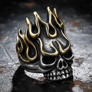 May include: A silver-toned skull ring with gold-colored flames. The ring features a detailed skull design with open eye sockets and a toothy grin. The flames are stylized and add a bold, edgy look to the jewelry.