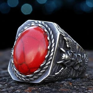 May include: A silver-toned ring with a large, oval, red stone. The ring has a detailed dragon design on the sides and a rope-like border around the stone. The ring is set against a dark background.