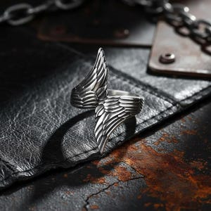 May include: A silver-colored ring with a wing design. The ring features detailed feather textures, with one wing extending upwards and the other wrapping around the finger. The ring is set against a dark, textured background.