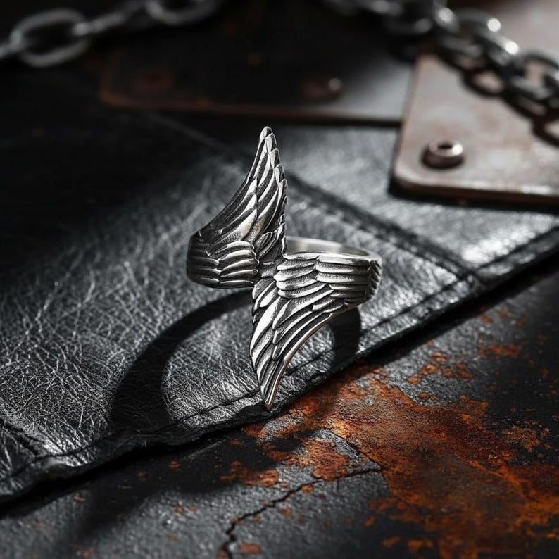 May include: A silver-colored ring with a wing design. The ring features detailed feather textures, with one wing extending upwards and the other wrapping around the finger. The ring is set against a dark, textured background.