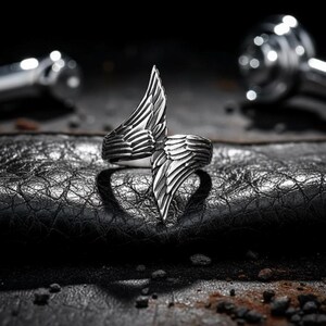 May include: A silver-colored ring with a wing design. The ring features two stylized wings that meet in the center, creating a bold and unique look. The wings are detailed with feather-like textures, adding depth and dimension to the jewelry.