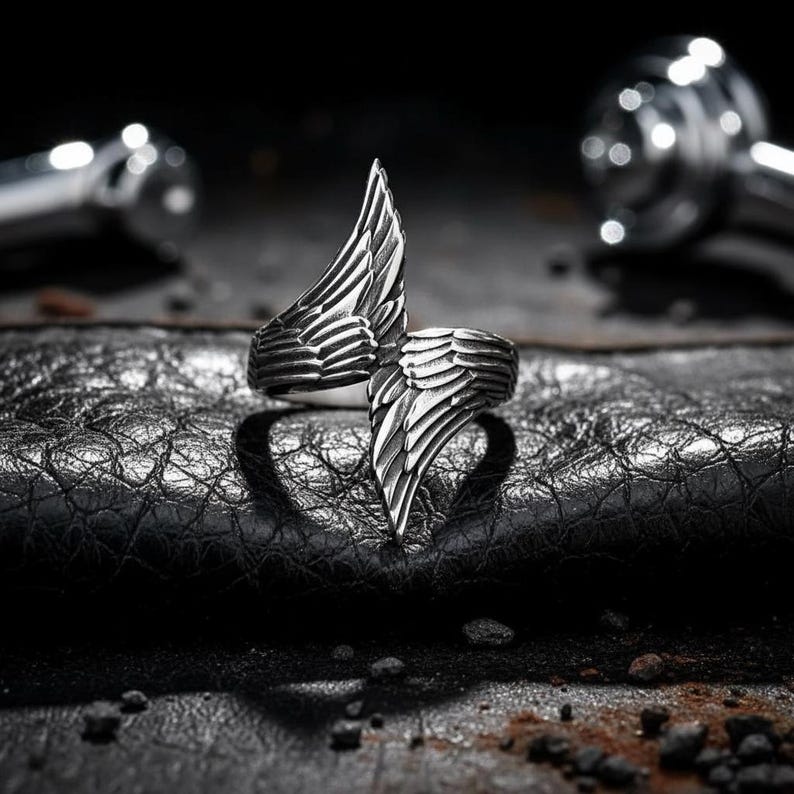 May include: A silver-colored ring with a wing design. The ring features two stylized wings that meet in the center, creating a bold and unique look. The wings are detailed with feather-like textures, adding depth and dimension to the jewelry.
