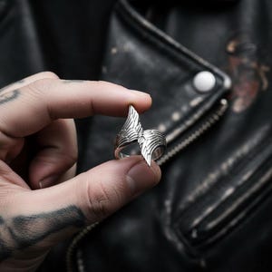 May include: A silver-colored ring with a wing design. The ring features detailed feather-like patterns and is held between two fingers. The background is a black leather jacket.
