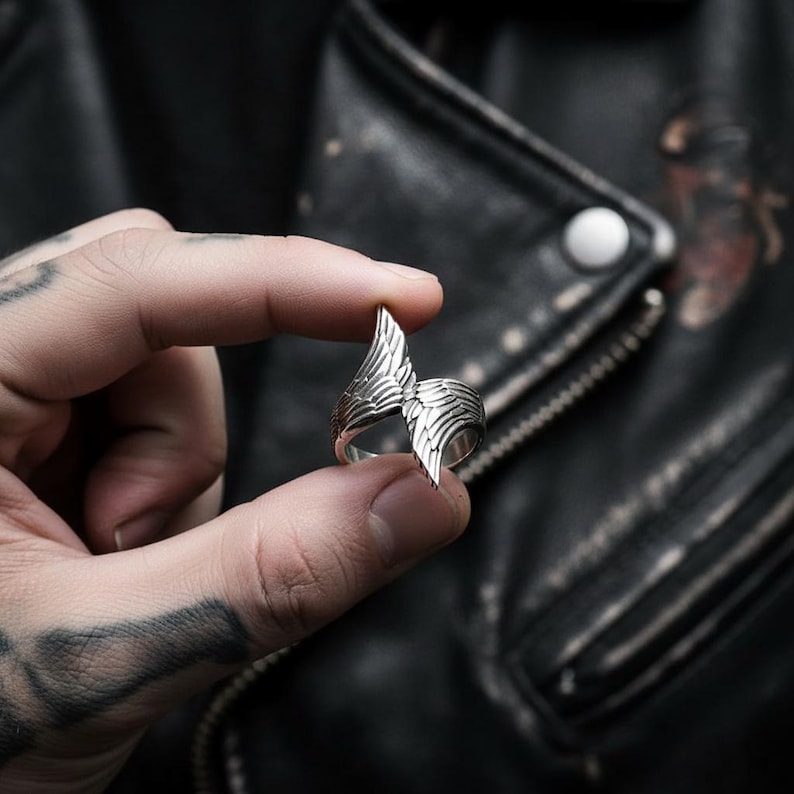 May include: A silver-colored ring with a wing design. The ring features detailed feather-like patterns and is held between two fingers. The background is a black leather jacket.