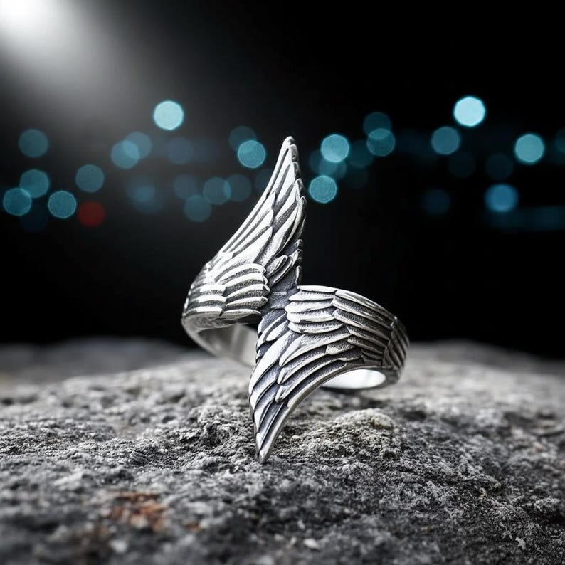 May include: A silver-colored ring with a wing design. The ring features detailed feather textures, with one wing extending upwards and the other curving downwards. The ring is set against a dark background with blurred lights, highlighting the jewelry's intricate design.