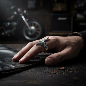 May include: A silver ring with a wing design. The ring is on a person's finger, with a motorcycle and amplifier in the background. The ring has a detailed, textured appearance.