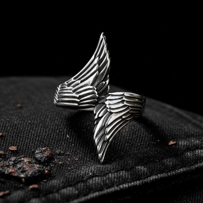May include: A silver-colored ring with a detailed wing design. The ring features two stylized wings that wrap around the finger, creating a bold and unique accessory. The ring is set against a dark background.