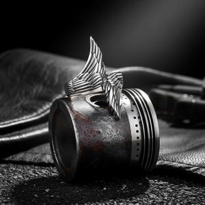 May include: A silver-colored ring with a detailed wing design. The ring is set against a dark background, resting on a metallic object. The wing design is the focal point, showcasing intricate feather detailing.