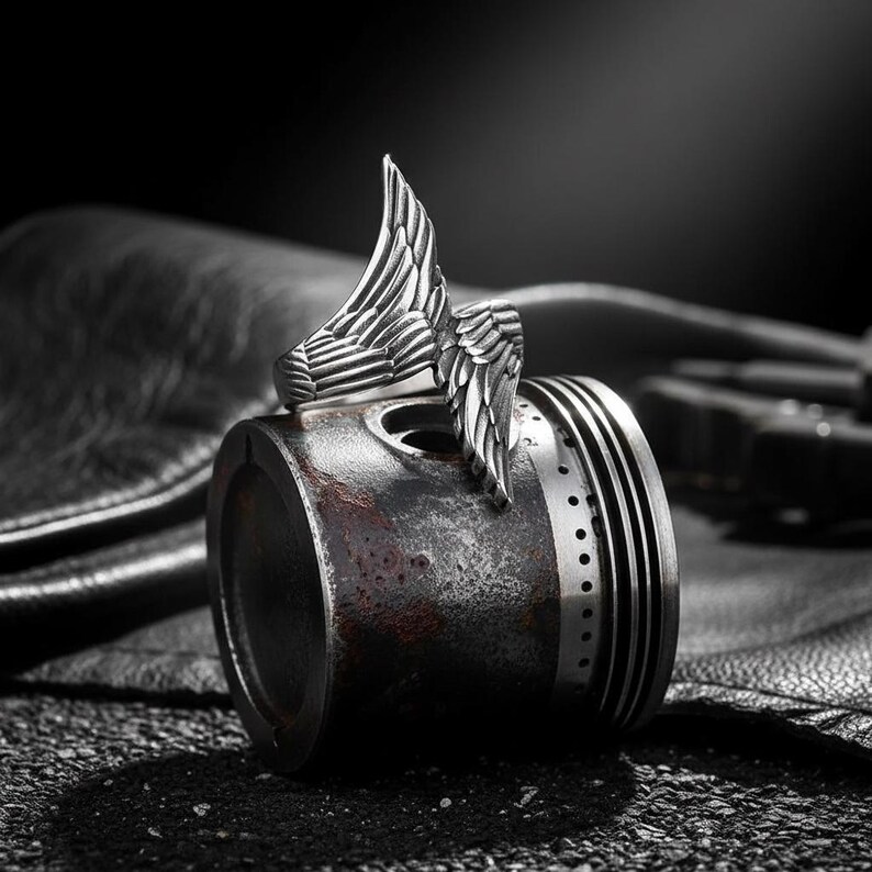 May include: A silver-colored ring with a detailed wing design. The ring is set against a dark background, resting on a metallic object. The wing design is the focal point, showcasing intricate feather detailing.