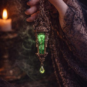May include: A bronze-colored pendant necklace with a rectangular green gemstone encased in an ornate frame. A teardrop-shaped green gem hangs below. The necklace is held against a dark background with a lit candle and smoky effect.