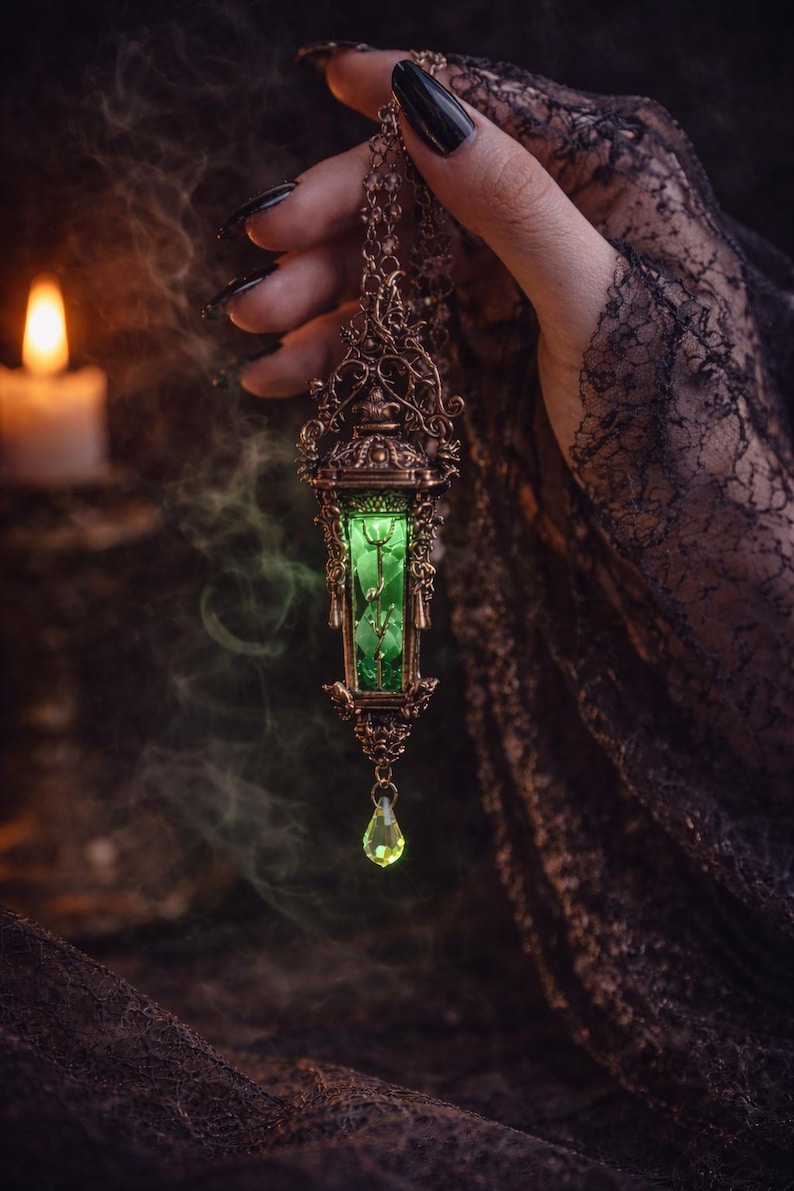 May include: A bronze-colored pendant necklace with a rectangular green gemstone encased in an ornate frame. A teardrop-shaped green gem hangs below. The necklace is held against a dark background with a lit candle and smoky effect.