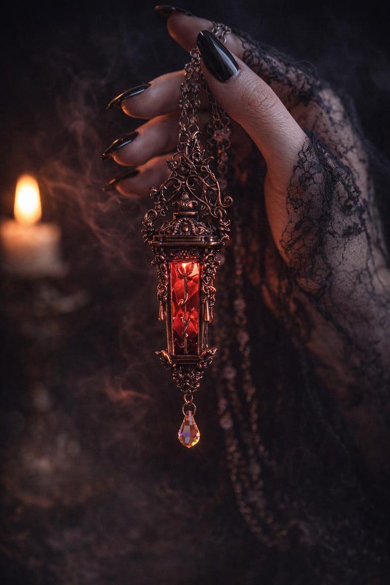 May include: Ornate bronze pendant necklace with a red interior, held against a dark background. The pendant has intricate filigree detailing and a teardrop-shaped crystal. The hand holding the necklace has long, dark nails and is wearing a black lace glove.