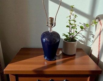 May include: A navy blue ceramic table lamp with a silver neck and two curved metal arms. The lamp sits on a wooden nightstand next to a small potted plant with white flowers. The lamp is plugged into a wall outlet.