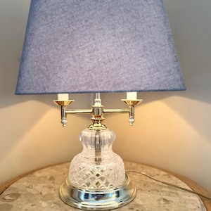 May include: A table lamp with a blue fabric shade. The lamp has a clear, cut-glass base with a gold-coloured metal accent and a silver-coloured base. The lamp has two light bulb holders.