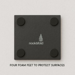Puede incluir: Un posavasos cuadrado negro con cuatro patas redondas de espuma negra. El posavasos tiene el texto "nook&fold" y un gr&aacute;fico de &aacute;rbol. La parte inferior del posavasos dice "FOUR FOAM FEET TO PROTECT SURFACES".