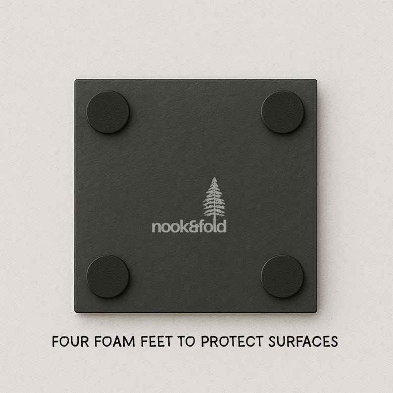 Puede incluir: Un posavasos cuadrado negro con cuatro patas redondas de espuma negra. El posavasos tiene el texto "nook&fold" y un gr&aacute;fico de &aacute;rbol. La parte inferior del posavasos dice "FOUR FOAM FEET TO PROTECT SURFACES".