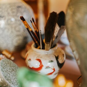 May include: A collection of paintbrushes and tools in a small, decorative ceramic jar. The jar is white with red and black painted designs. Various paintbrushes with gold and brown handles are visible, along with a large brush and a palette knife.