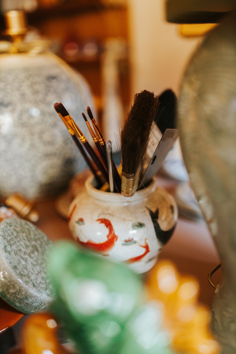 May include: A collection of paintbrushes and tools in a small, decorative ceramic jar. The jar is white with red and black painted designs. Various paintbrushes with gold and brown handles are visible, along with a large brush and a palette knife.