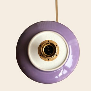 May include: Overhead view of a purple and white ceramic light fixture. The fixture has a circular design with a brass-colored socket in the center. A beige cord extends from the top.