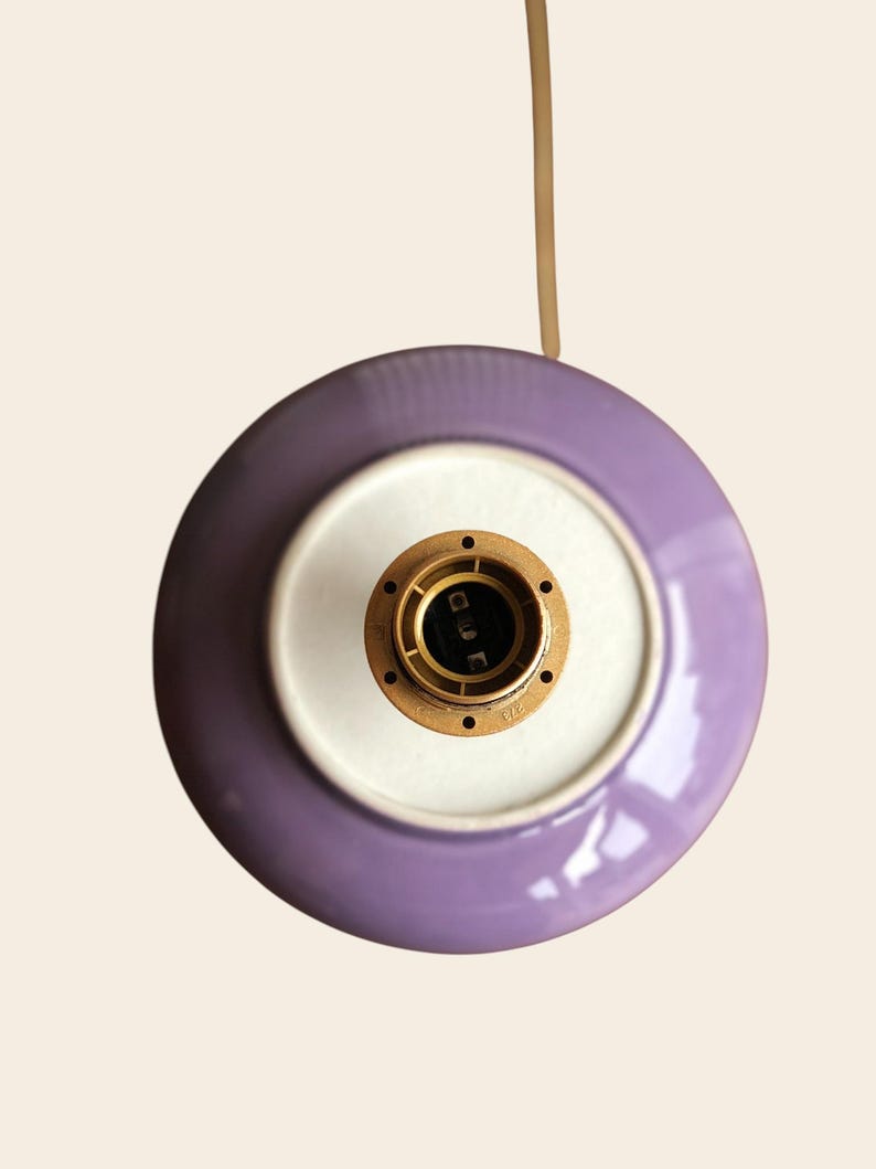 May include: Overhead view of a purple and white ceramic light fixture. The fixture has a circular design with a brass-colored socket in the center. A beige cord extends from the top.