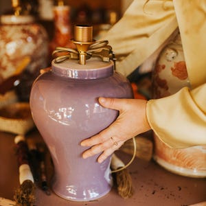 May include: A lavender ceramic lamp base with a gold-toned socket and cord. The lamp has a rounded shape and is being held on a pink table. Other decorative lamps are visible in the background.