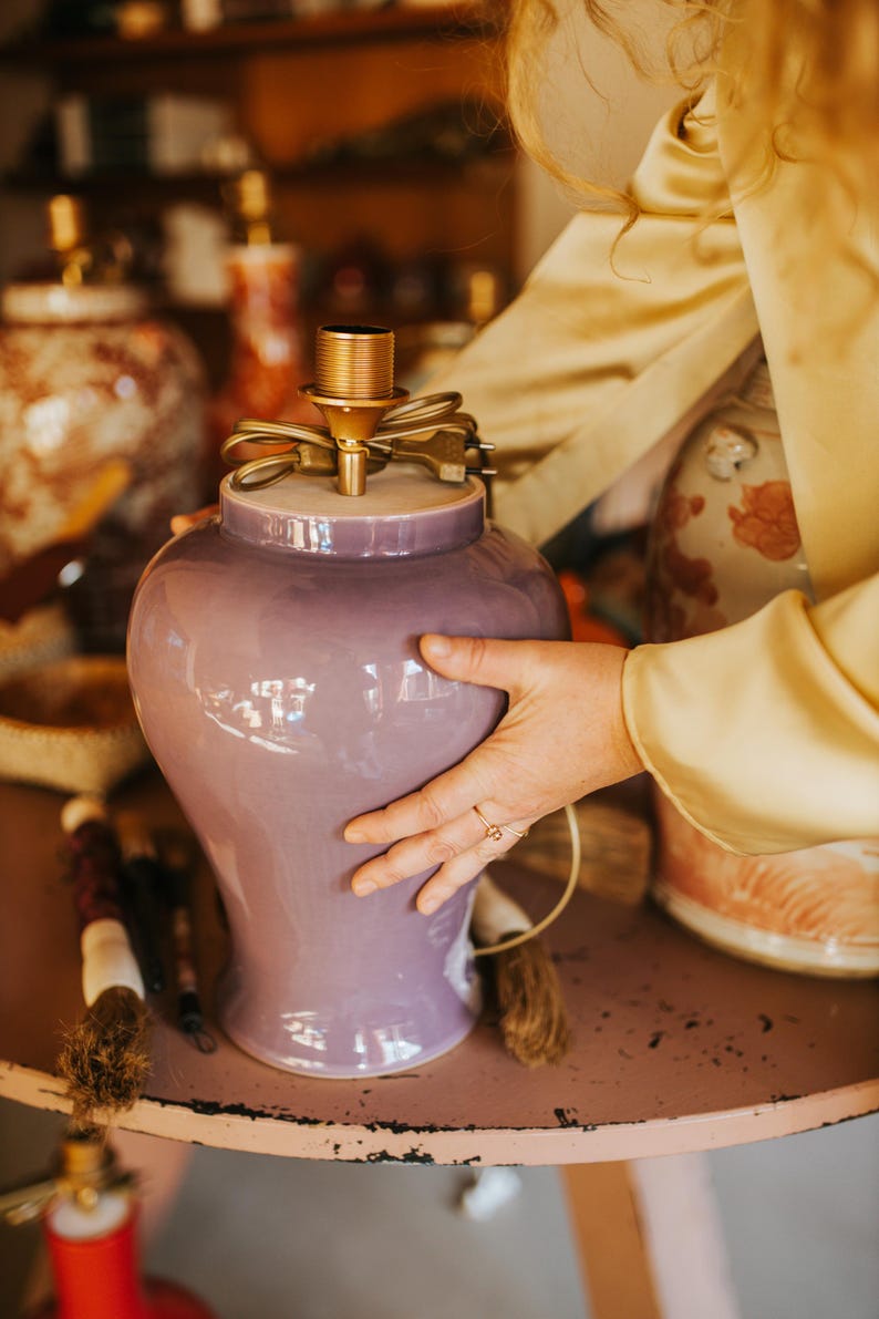May include: A lavender ceramic lamp base with a gold-toned socket and cord. The lamp has a rounded shape and is being held on a pink table. Other decorative lamps are visible in the background.