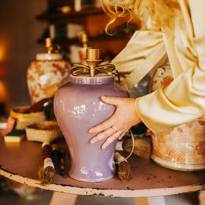 May include: A lavender ceramic lamp with a gold-colored top and a decorative bow. The lamp is on a pink table, with other decorative items in the background. The lamp is being held by a person.