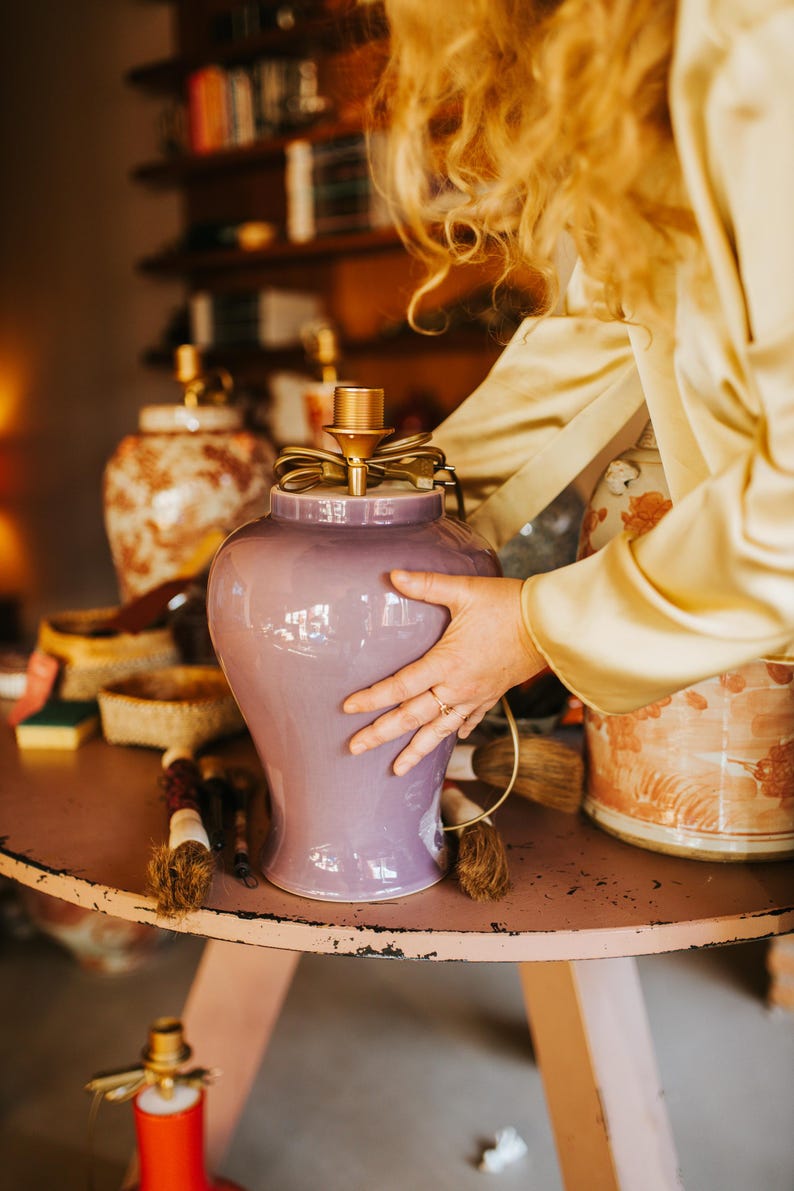 May include: A lavender ceramic lamp with a gold-colored top and a decorative bow. The lamp is on a pink table, with other decorative items in the background. The lamp is being held by a person.